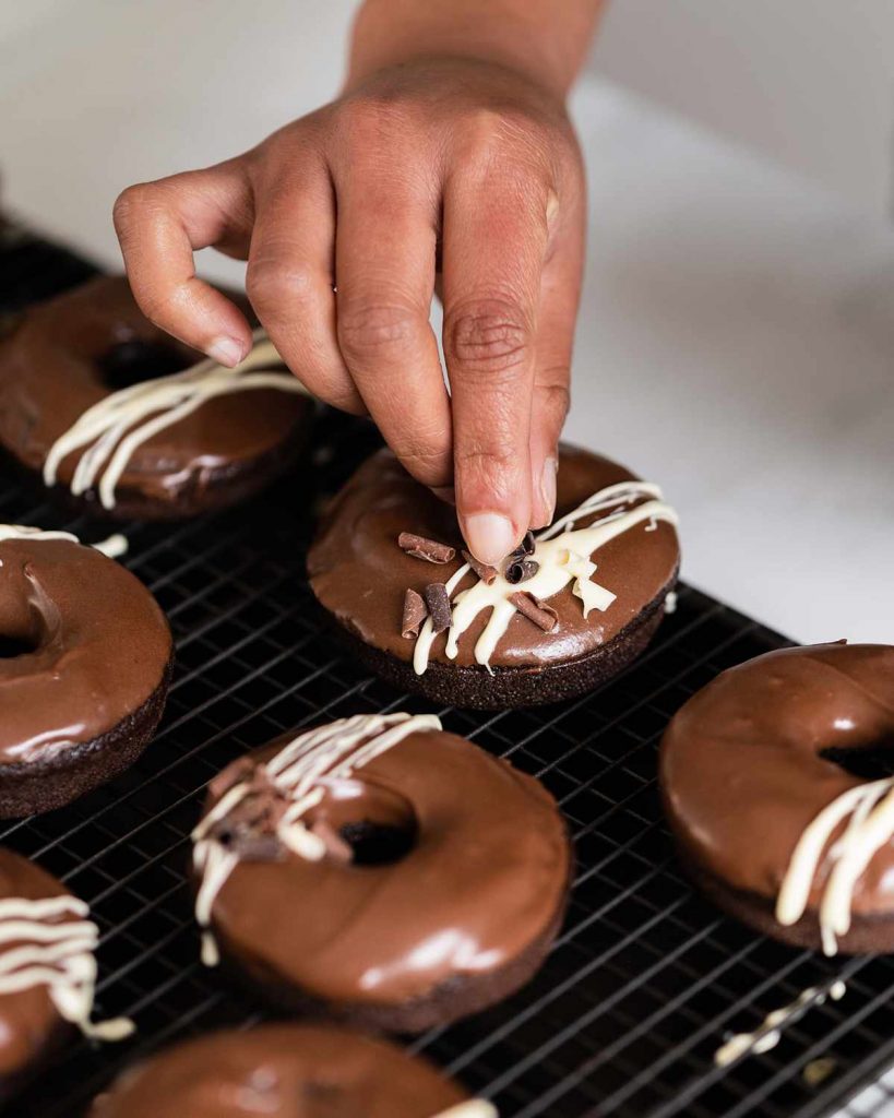 Adding the finishing touches to the chocolate fudge doughnuts: a white chocolate drizzle and some chocolate curls. Recipe by movers and bakers