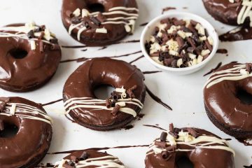 Soft, light and moist chocolate doughnuts with a delicious chocolate fudge glaze, white chocolate drizzle and chocolate curls to finish. Recipe by movers and bakers