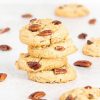 A stack of four coconut pecan cookies is the focus, with scattered pecans and additional cookies dotted around. Recipe by movers and bakers