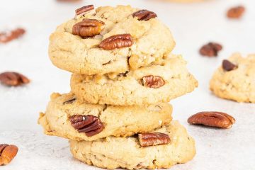 A stack of four coconut pecan cookies is the focus, with scattered pecans and additional cookies dotted around. Recipe by movers and bakers