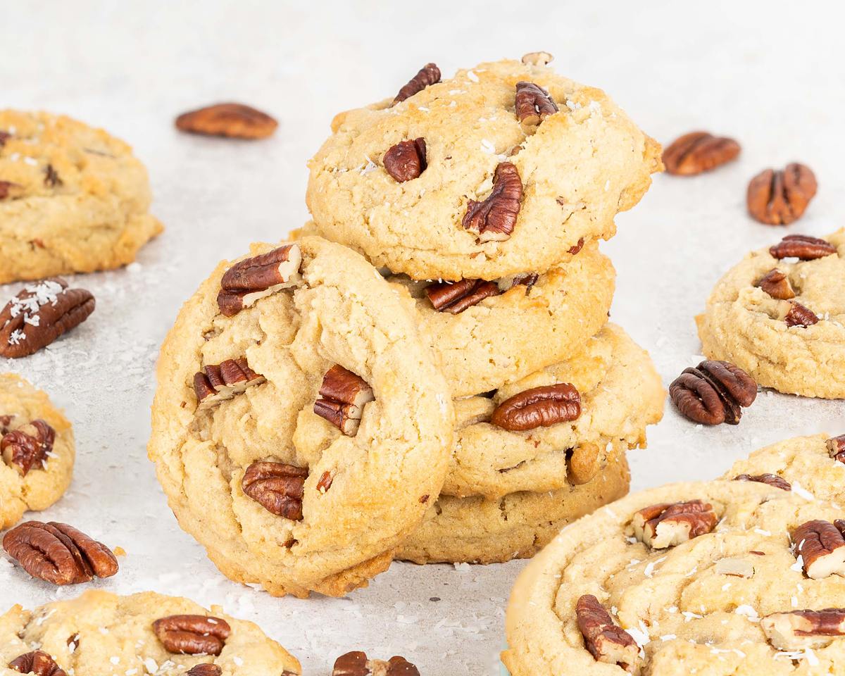 A stack of four coconut pecan cookies is the focus, with scattered pecans and additional cookies dotted around. Recipe by movers and bakers