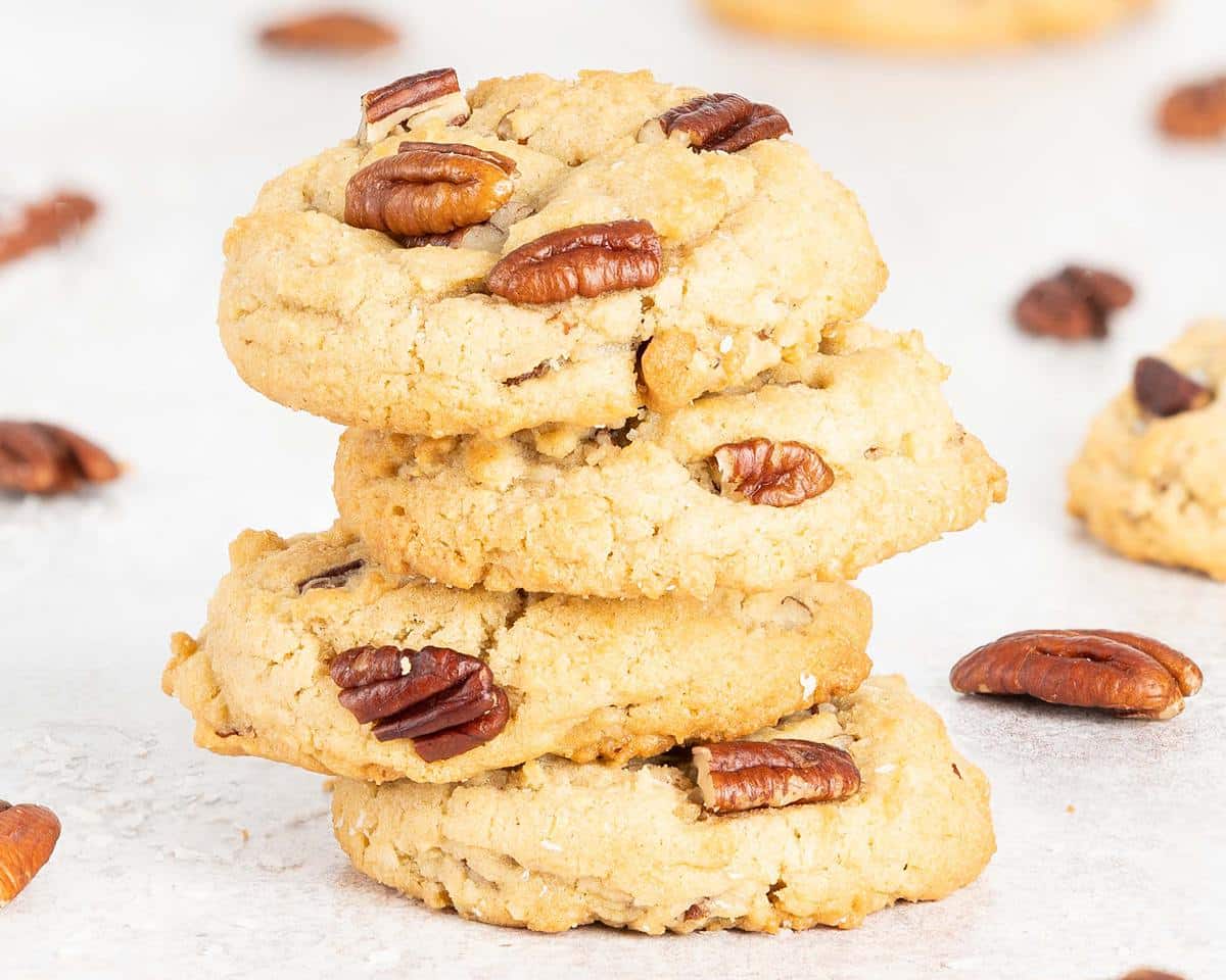 A stack of four coconut pecan cookies is the focus, with scattered pecans and additional cookies dotted around. Recipe by movers and bakers