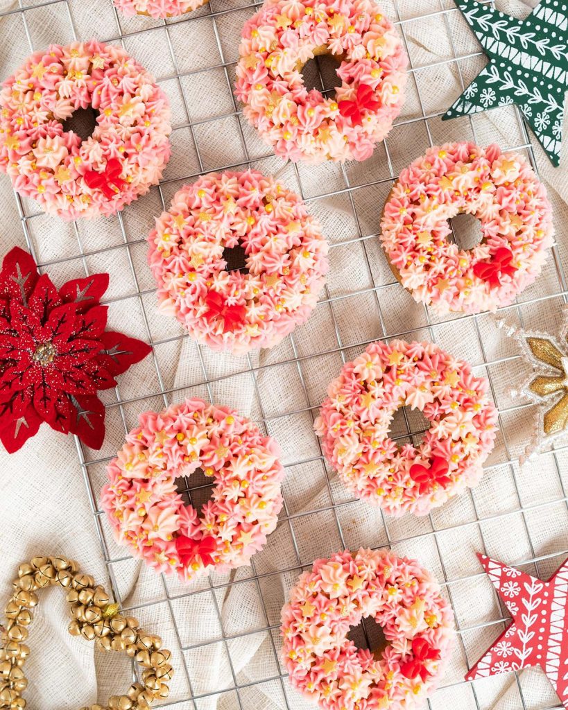 Christmas wreath doughnuts. Light and fluffy vanilla doughnuts topped with a light vanilla buttercream in pretty shades of pink. Finished with some gorgeous delicate gold sprinkles and a red fondant bow and you have a showstopper doughnut! Recipe by movers and bakers