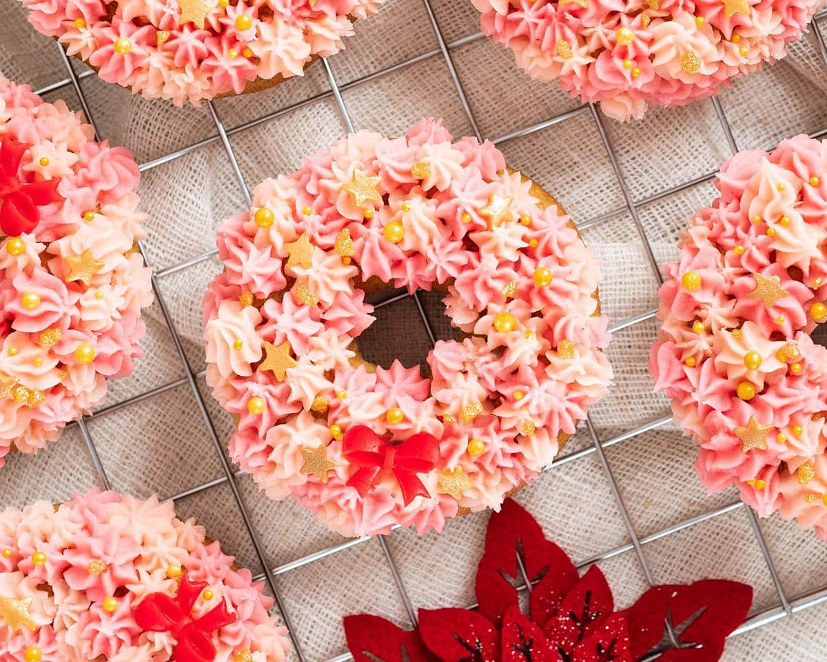 Christmas wreath doughnuts. Light and fluffy vanilla doughnuts topped with a light vanilla buttercream in pretty shades of pink. Finished with some gorgeous delicate gold sprinkles and a red fondant bow and you have a showstopper doughnut! Recipe by movers and bakers