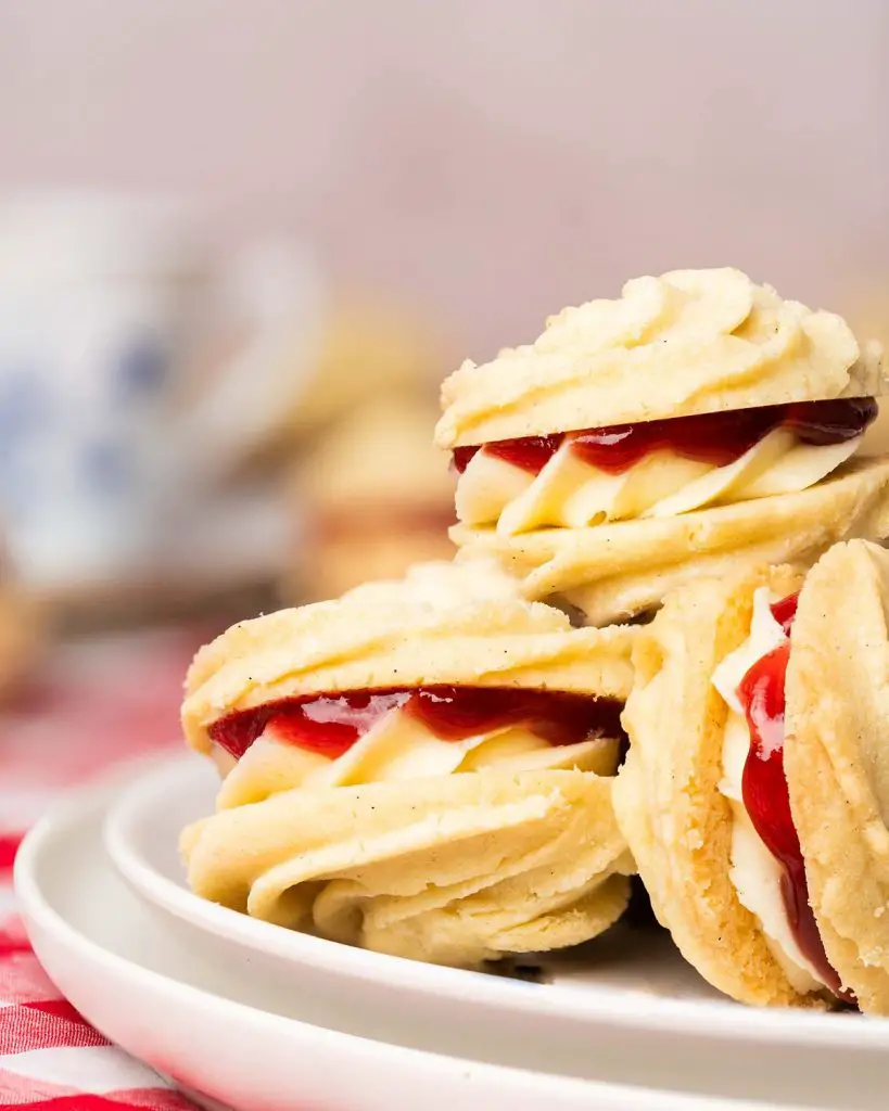 Viennese whirls are a delightful British sandwich biscuit! Two buttery piped shortbread biscuits are filled with soft vanilla buttercream and raspberry jam, making the perfect tea time treat! Recipe by movers and bakers