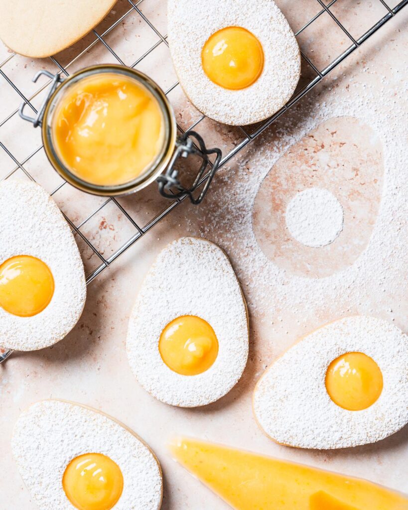 Easter egg lemon curd cookies. Two egg sugar cookies sandwiched together with lemon curd, dusted with icing sugar. The top cookie has a small circular hole cut from its centre, allowing the golden yellow lemon curd filling to peek through, mimicking the look of a fried egg. Recipe by movers and bakers