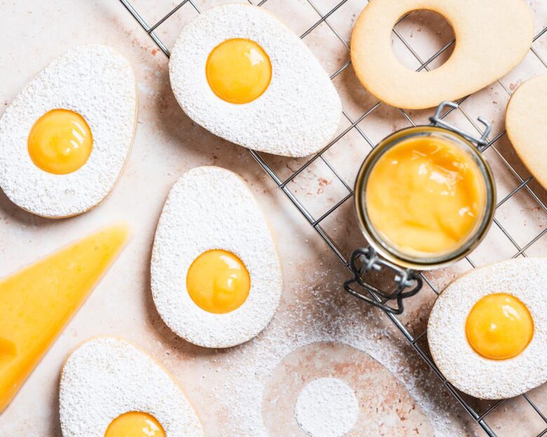 Easter egg lemon curd cookies. Two egg sugar cookies sandwiched together with lemon curd, dusted with icing sugar. The top cookie has a small circular hole cut from its centre, allowing the golden yellow lemon curd filling to peek through, mimicking the look of a fried egg. Recipe by movers and bakers