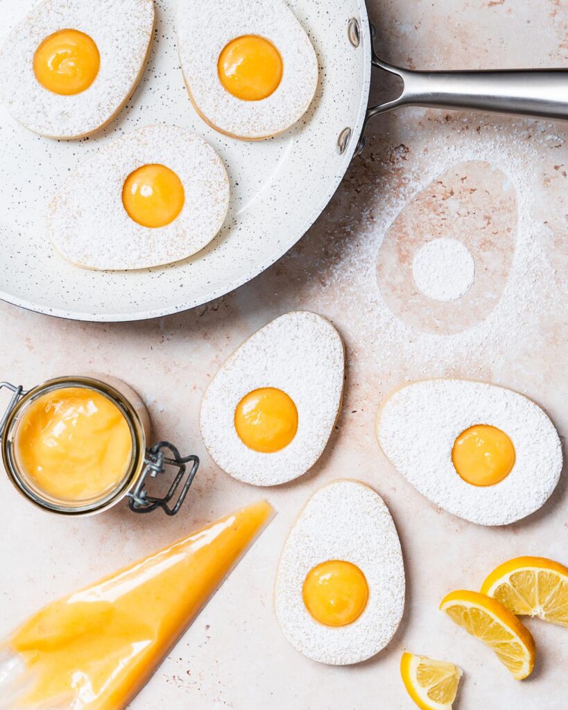 Easter egg lemon curd cookies. Two egg sugar cookies sandwiched together with lemon curd, dusted with icing sugar. The top cookie has a small circular hole cut from its centre, allowing the golden yellow lemon curd filling to peek through, mimicking the look of a fried egg. Recipe by movers and bakers