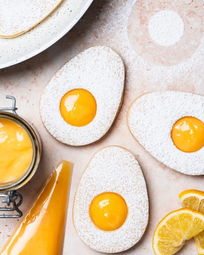 Easter egg lemon curd sugar cookies. Two egg sugar cookies sandwiched together with lemon curd, dusted with icing sugar. The top cookie has a small circular hole cut from its centre, allowing the golden yellow lemon curd filling to peek through, mimicking the look of a fried egg. Recipe by movers and bakers