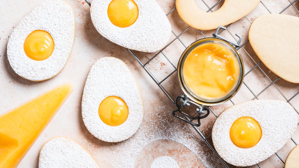 Easter egg lemon curd cookies. Two egg sugar cookies sandwiched together with lemon curd, dusted with icing sugar. The top cookie has a small circular hole cut from its centre, allowing the golden yellow lemon curd filling to peek through, mimicking the look of a fried egg. Recipe by movers and bakers
