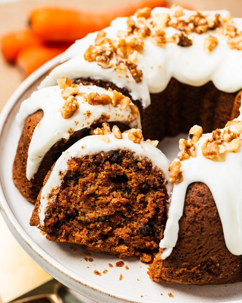 Carrot bundt cake on a cake stand with cream cheese glaze dripping into the ridges, topped with chopped walnuts. Rich, moist, perfectly spiced and totally irresistible! Recipe by movers and bakers
