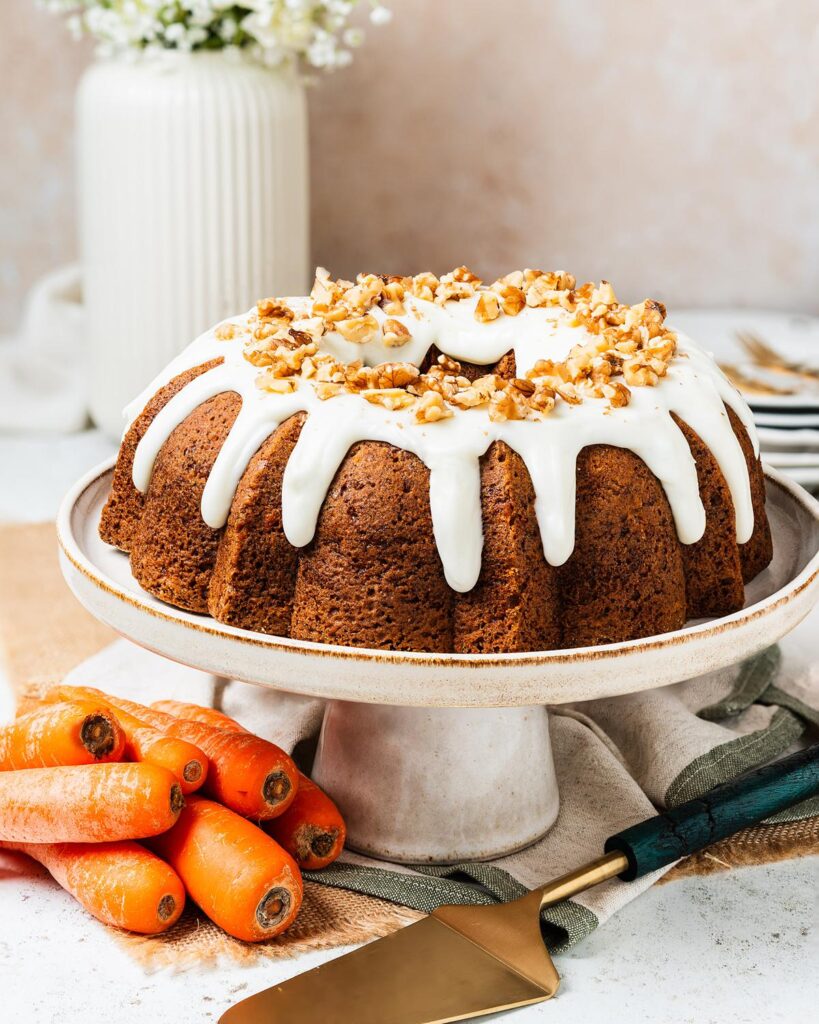 Carrot bundt cake on a cake stand with cream cheese glaze dripping into the ridges, topped with chopped walnuts. Rich, moist, perfectly spiced and totally irresistible! Recipe by movers and bakers