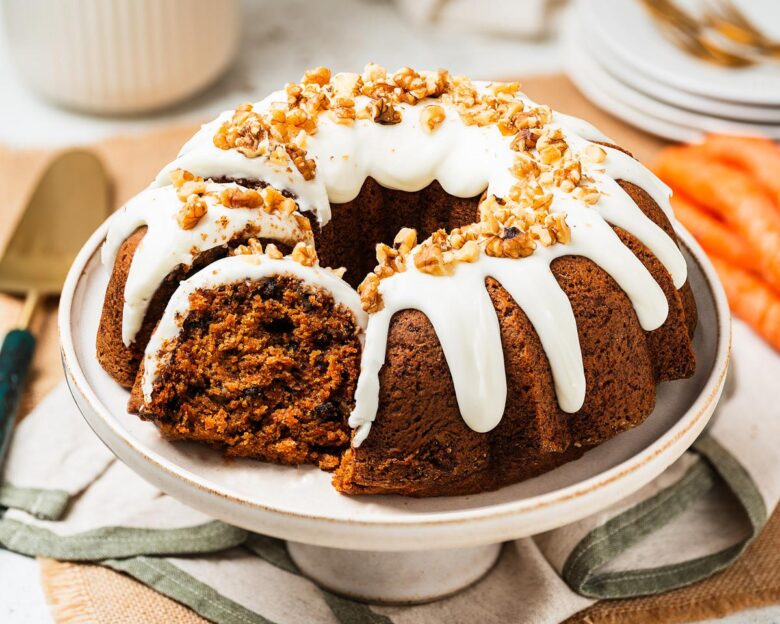 Carrot bundt cake on a cake stand with cream cheese glaze dripping into the ridges, topped with chopped walnuts. Rich, moist, perfectly spiced and totally irresistible! Recipe by movers and bakers