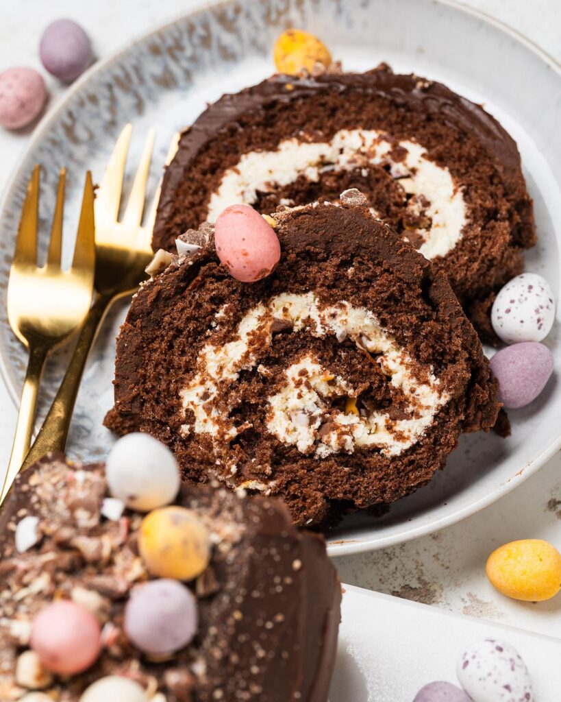 Mini Egg Swiss Roll Cake sliced to show cream filling and chopped mini eggs inside, topped with chocolate ganache and whole mini eggs. Recipe by movers and bakers