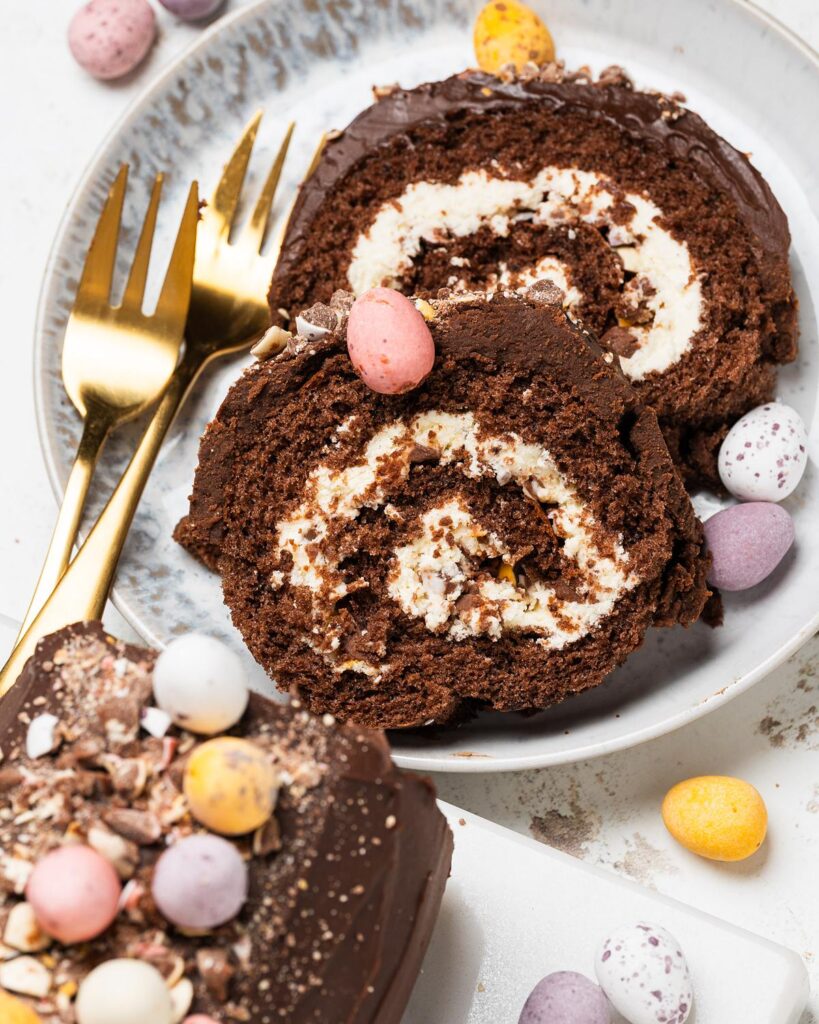 Mini Egg Swiss Roll Cake sliced to show cream filling and chopped mini eggs inside, topped with chocolate ganache and whole mini eggs. Recipe by movers and bakers