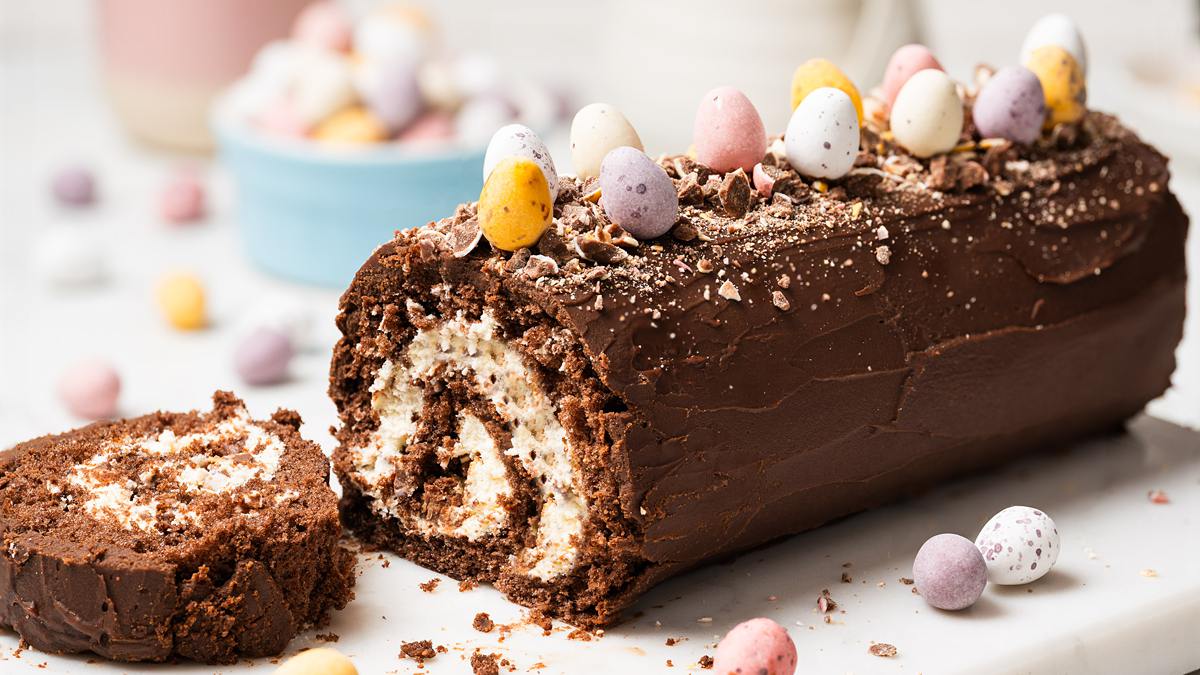 Mini Egg Swiss Roll Cake sliced to show cream filling and chopped mini eggs inside, topped with chocolate ganache and whole mini eggs. Recipe by movers and bakers