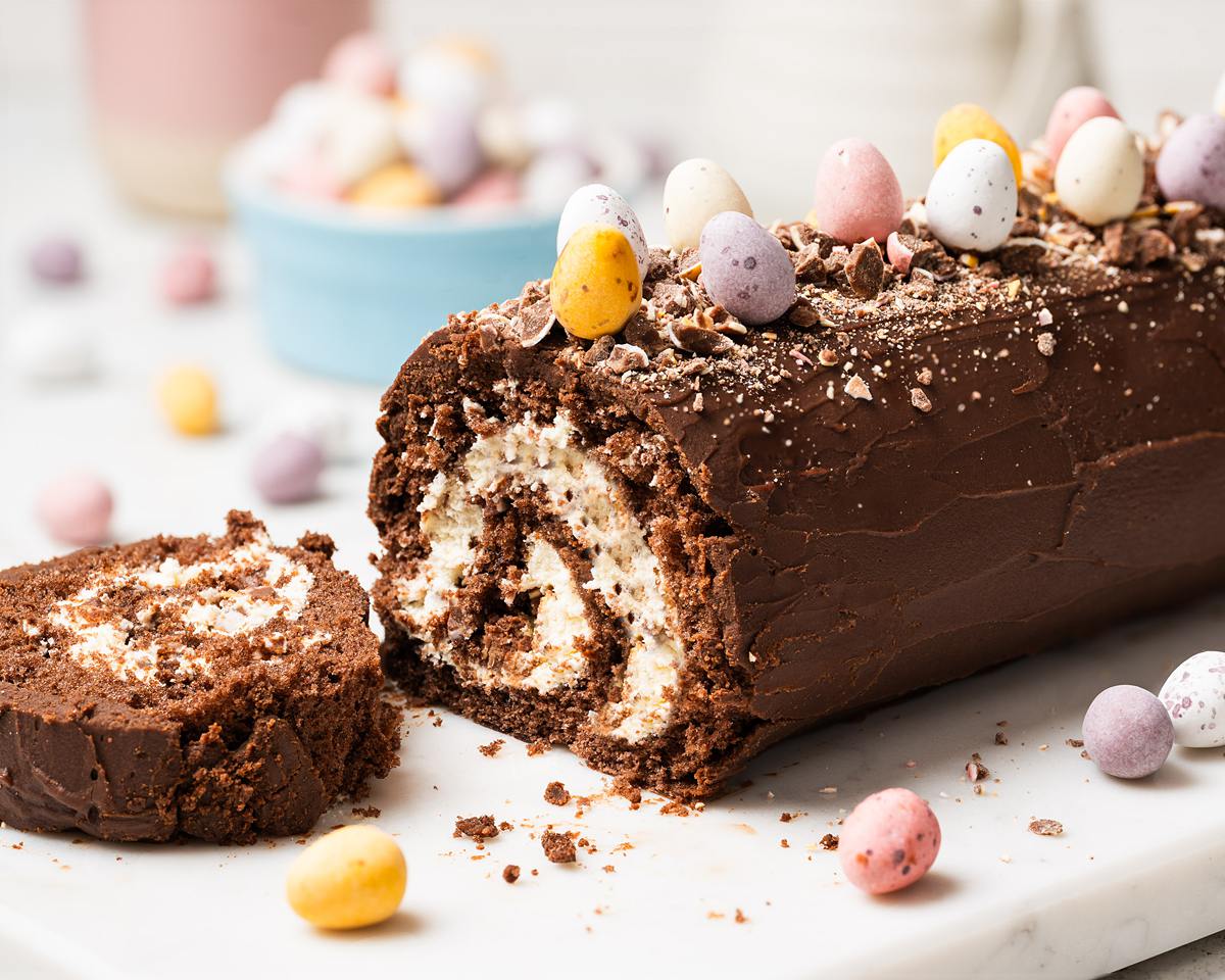 Mini Egg Swiss Roll Cake sliced to show cream filling and chopped mini eggs inside, topped with chocolate ganache and whole mini eggs. Recipe by movers and bakers