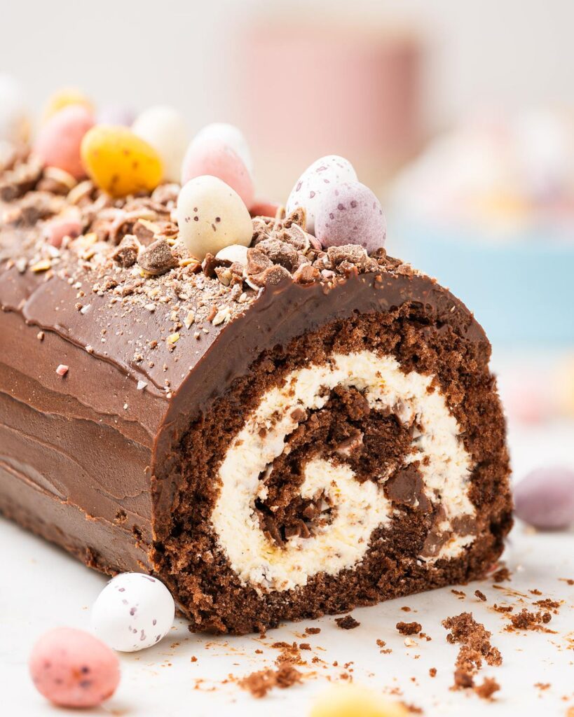 Mini Egg Swiss Roll Cake sliced to show cream filling and chopped mini eggs inside, topped with chocolate ganache and whole mini eggs. Recipe by movers and bakers
