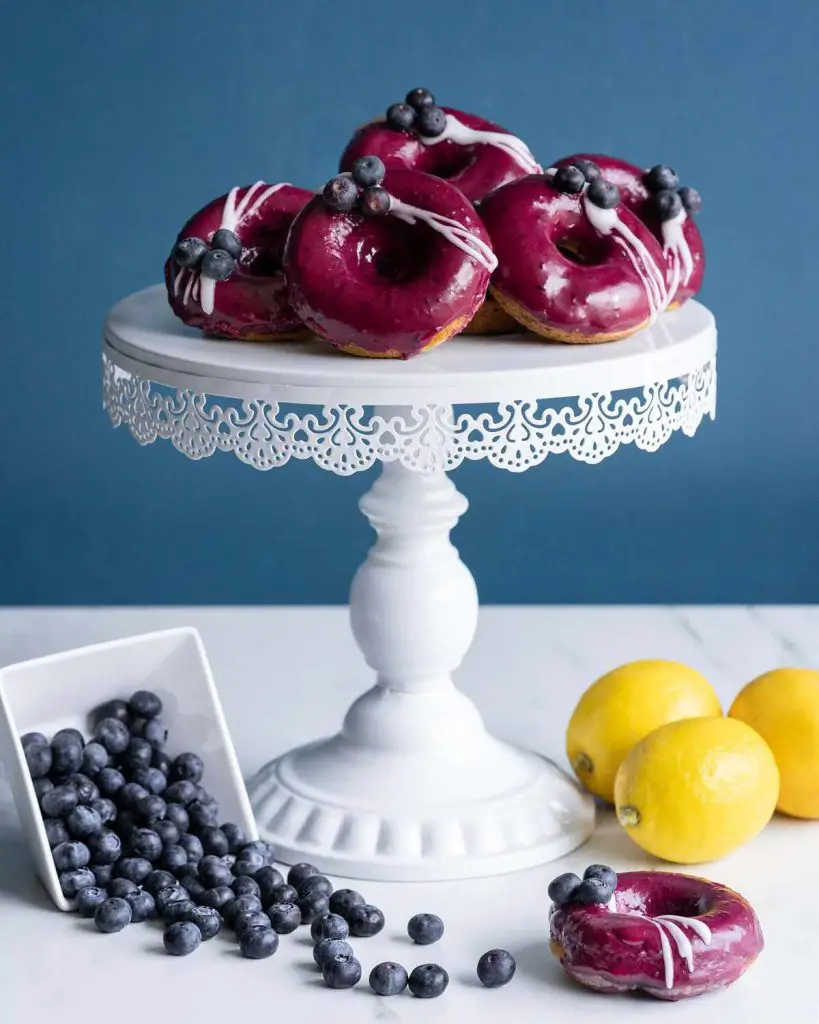 A pile of lemon blueberry donuts on a cake stand surrounded by blueberries and whole lemons. Stunning!