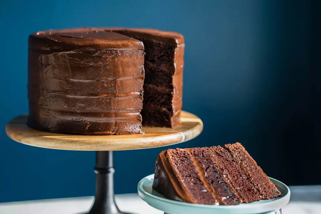 Rich and decadent four layer chocolate fudge cake on a stand with a slice cut out on a plate.