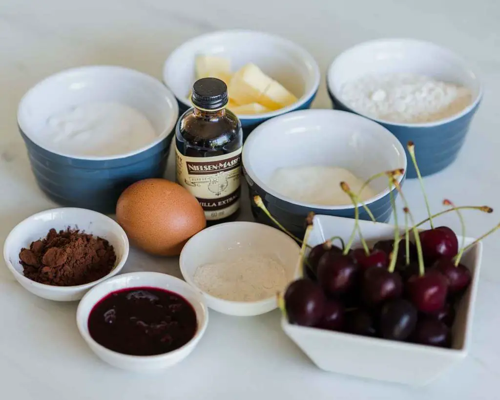 Ingredients needed for Black Forest cupcakes: plain flour, cocoa powder, baking powder, bicarbonate of soda (baking soda), salt, unsalted butter, caster sugar, egg, vanilla, yogurt, Kirsch, black cherry conserve, icing (powdered) sugar, double cream, fresh cherries and dark chocolate. Recipe by movers and bakers