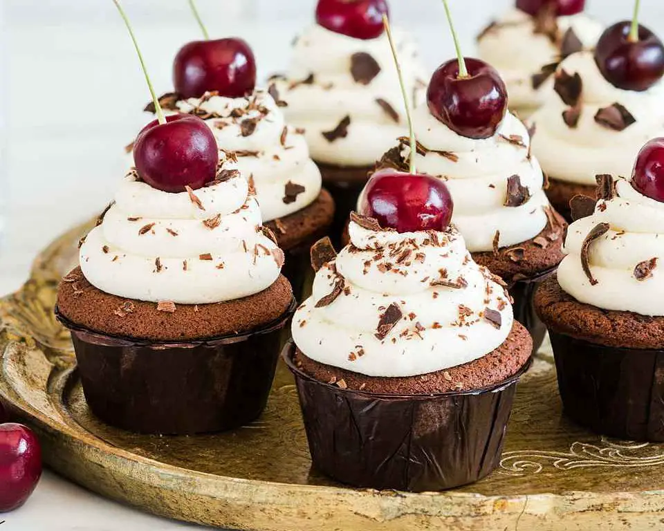 Black Forrest cupcakes are light but deeply chocolatey cupcakes, laced with Kirsch and filled with delicious black cherry compote, then topped with a buttercream swirl, grated chocolate and a cherry on top. Recipe by movers and bakers