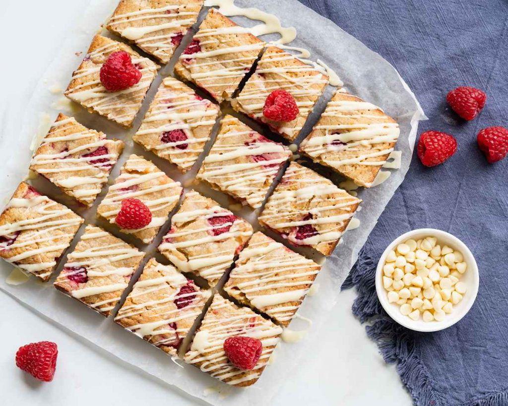 A batch of raspberry white chocolate blondies, cut into squares with additional raspberries and white chocolate chips for decoration. Recipe by movers and bakers