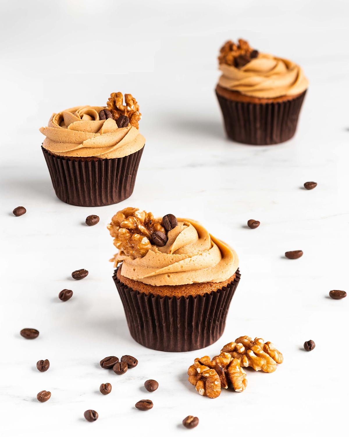 Three cupcakes staggered on a table with a sprinkling of coffee beans and walnuts surrounding them