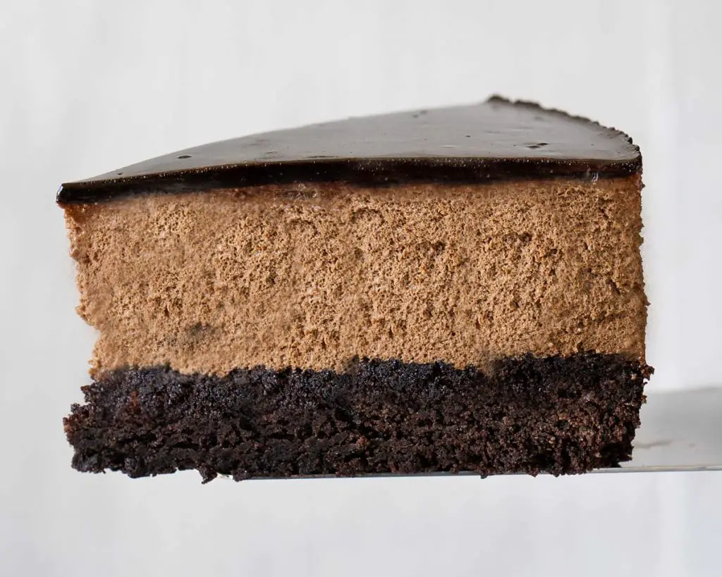 A slice of brownie mousse cake, showing the layers of brownie, mousse and glaze all in perfect proportion. This brownie mousse cake is rich and purely delightful! Recipe by movers and bakers