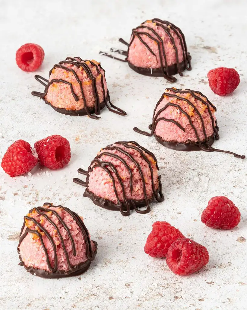 Raspberry and dark chocolate macaroons lay scattered with some raspberries placed around them. Recipe by movers and bakers