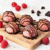Raspberry chocolate macaroons on a wooden serving board with scattered raspberries and dark chocolate around. Recipe by movers and bakers