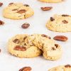 Coconut pecan cookies. Several cookies are laid across the table, scattered with desiccated coconut and pecan nuts. One in the middle has a bite taken out of it and leans up against another cookie. Recipe by movers and bakers